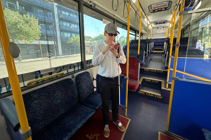 A woman wearing sunglasses and a white shirt stands on a bus using her mobile phone with her cane tucked under her arm. 