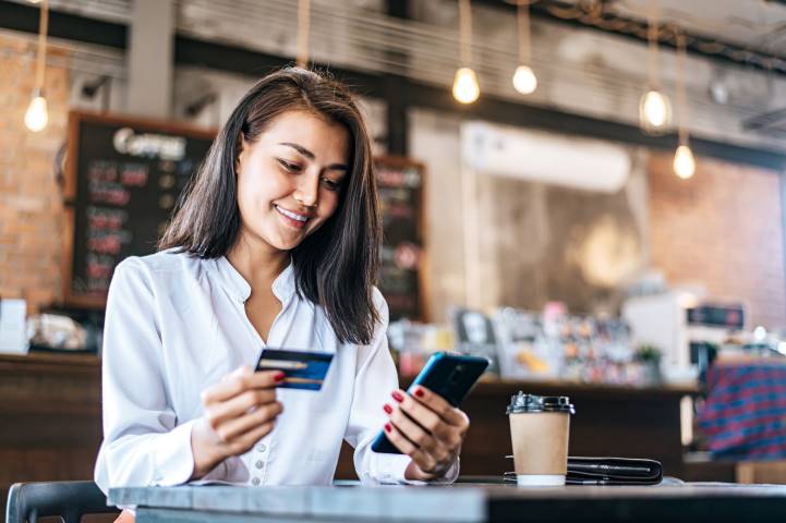 A smiling woman is sitting in a coffee shop, looking at her mobile phone. She is holding a credit card in her other hand.