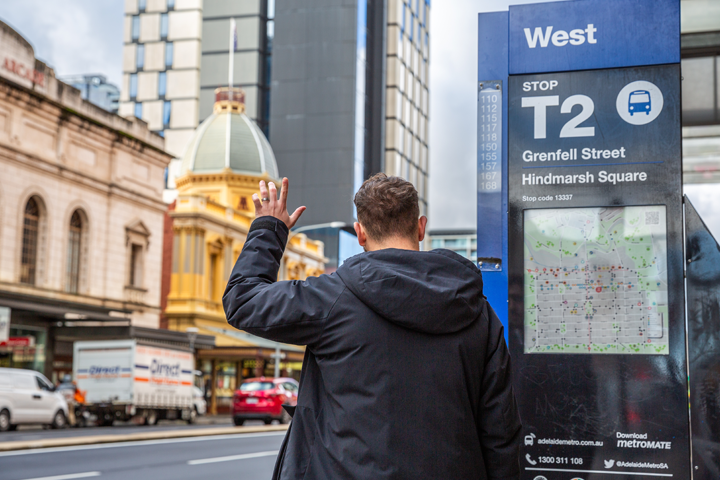 A passenger hailing a bus in the city at a bus stop/ Their left in the hand is in the air, which will hail the bus.